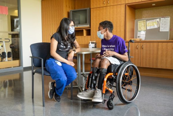 An adult on the left and a teenager sitting on wheelchair on the right