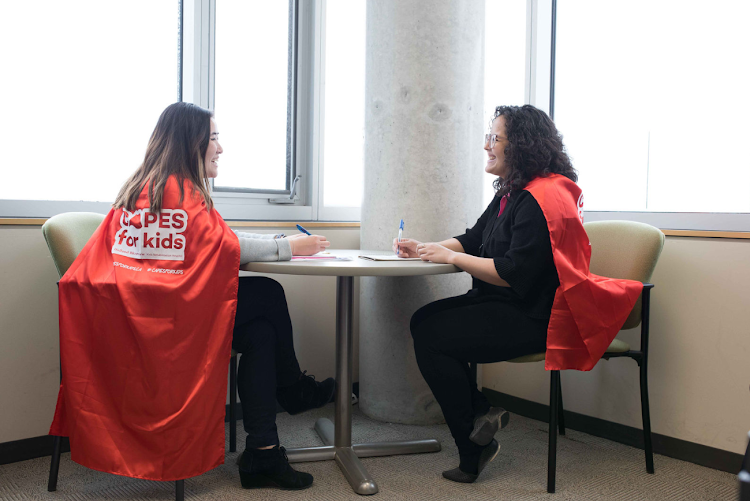 Two women wearing capes facing each other while sitting at a table. 