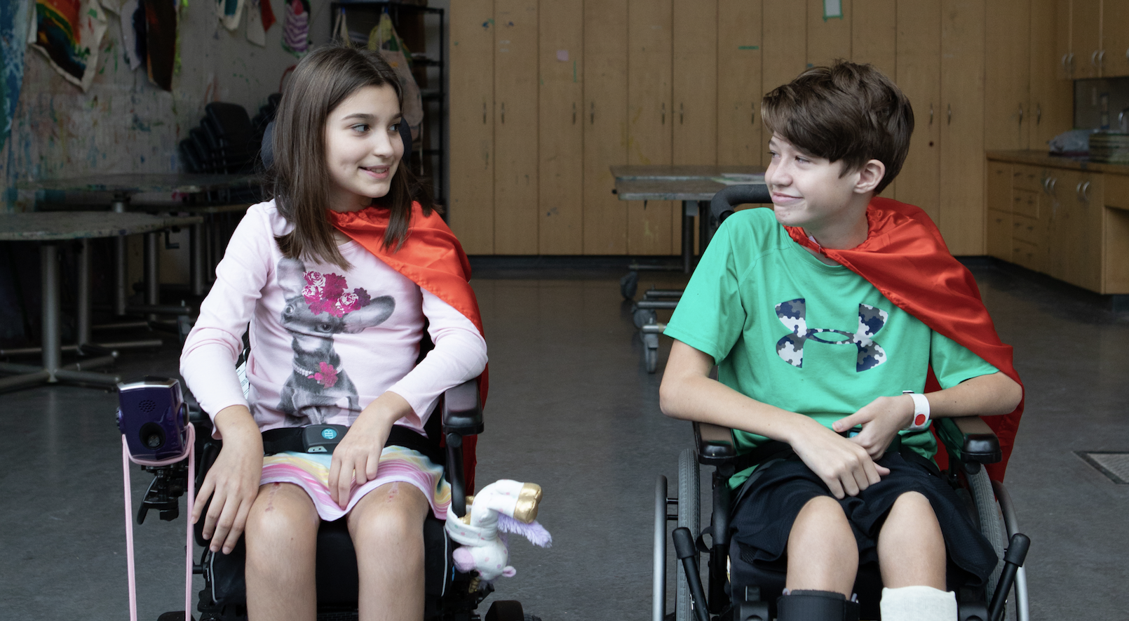 Two youth who use wheelchairs sitting together in an art studio wearing capes for Capes for Kids.