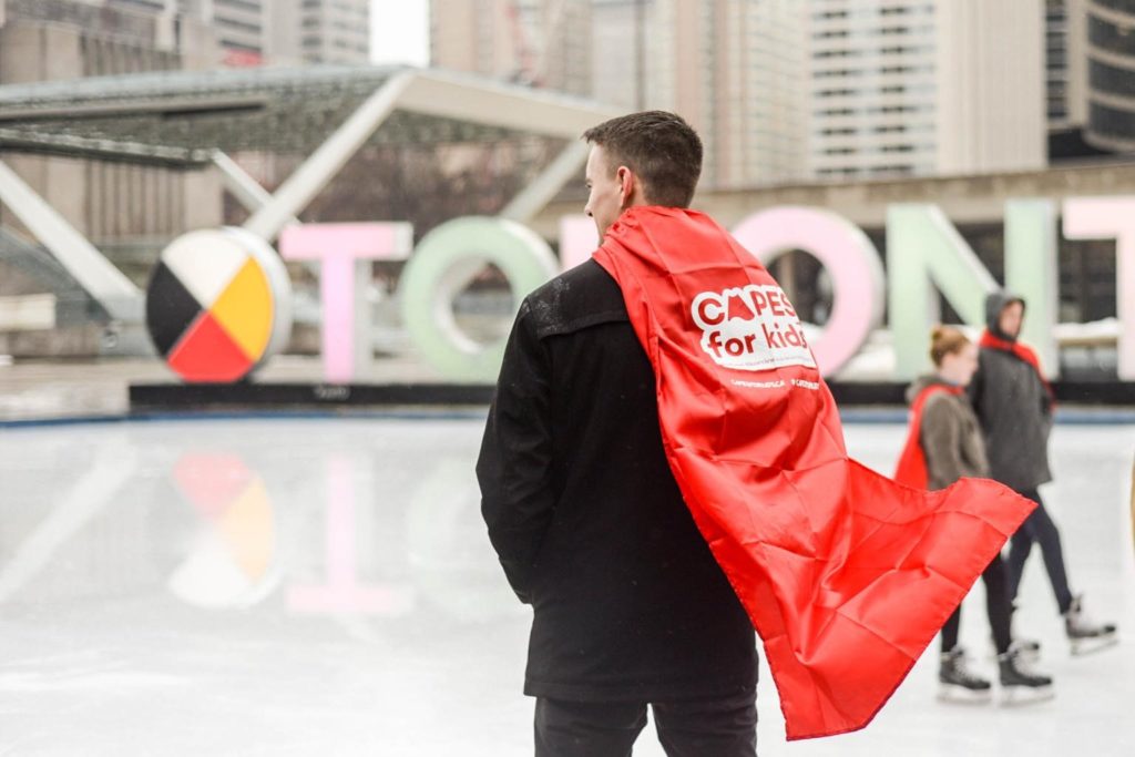 A man participating in corporate giving through Capes for Kids stands in front of Nathan Phillips Square. He is wearing a black coat and a red cape.