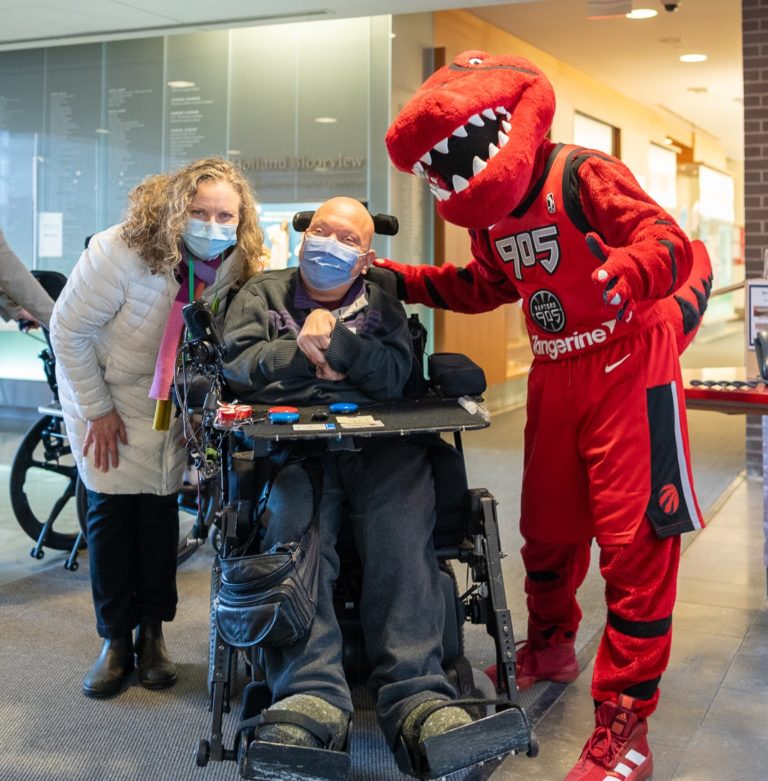 Two adults posing with the Raptors 905 dinosaur mascot in the Holland Bloorview lobby.
