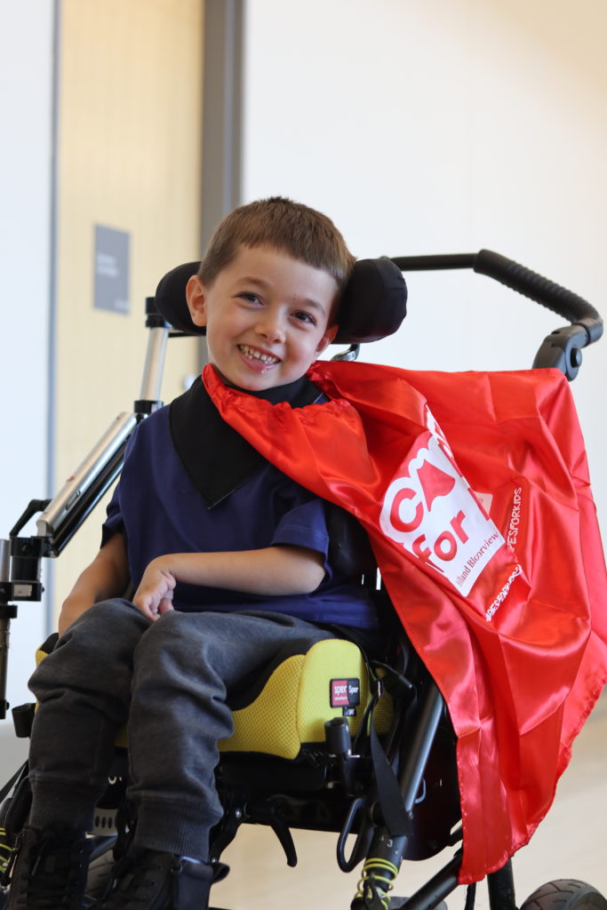 A young boy with light skin tone and short brown hair smiling and wearing a red cape. He is using a wheelchair.