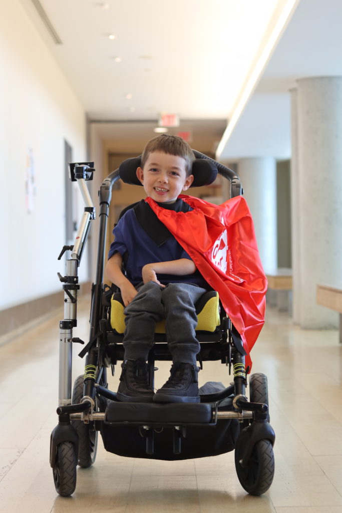 A young boy with light skin tone and short brown hair smiling and wearing a red cape. He is using a wheelchair.