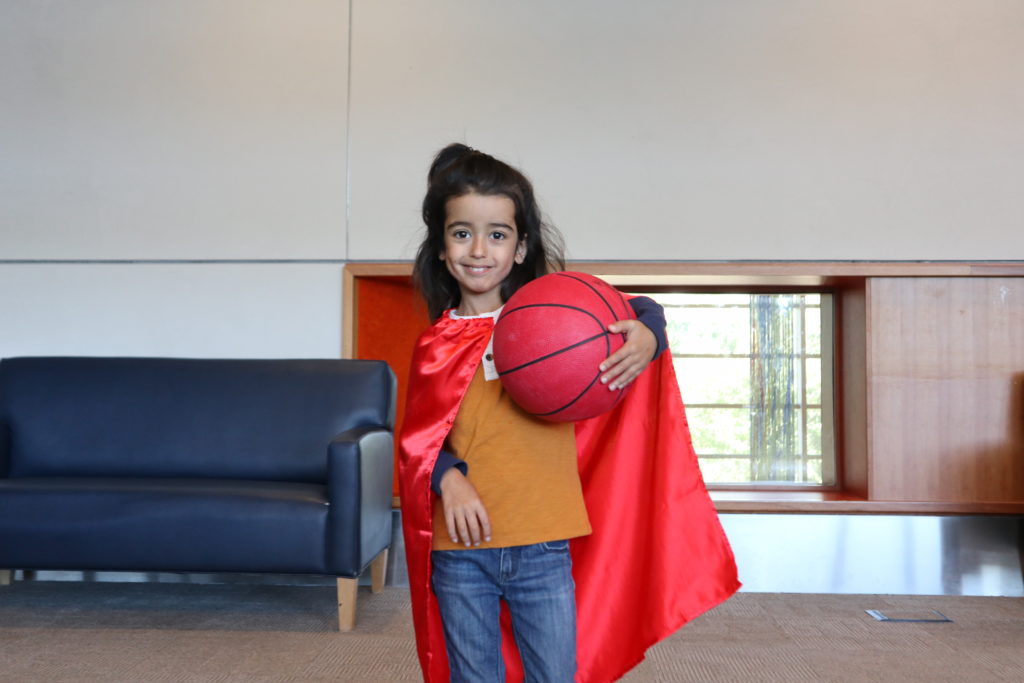 A boy with medium-light skin tone and long black hair wearing a red cape and holding a basketball.