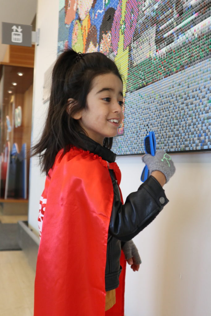 A boy with medium-light skin tone and long black hair wearing a red cape. He is looking at a piece of art on the wall.