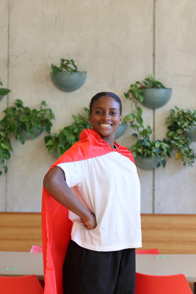 A teenager with medium-dark skin tone and black hair wearing a red cape and a white t-shirt. She is standing in a superhero pose with her fist on her hip.