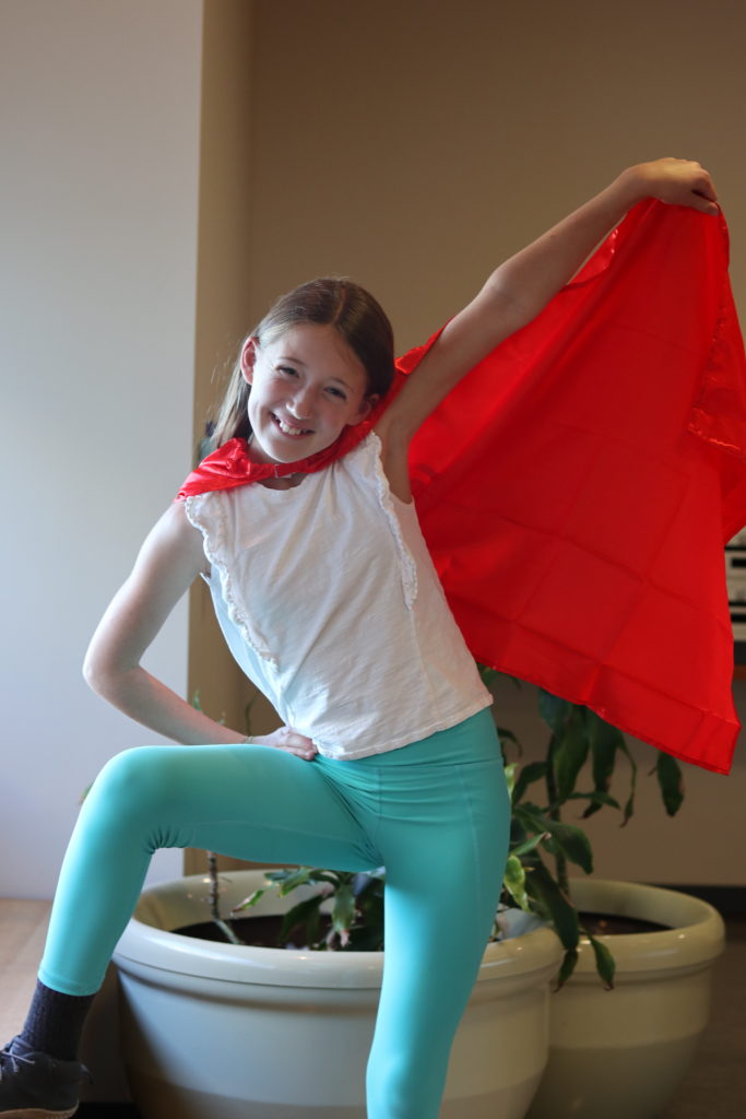 A young girl with light skin tone and long brown hair wearing a red cape. She is holding a part of her cape over her head in a superhero pose.