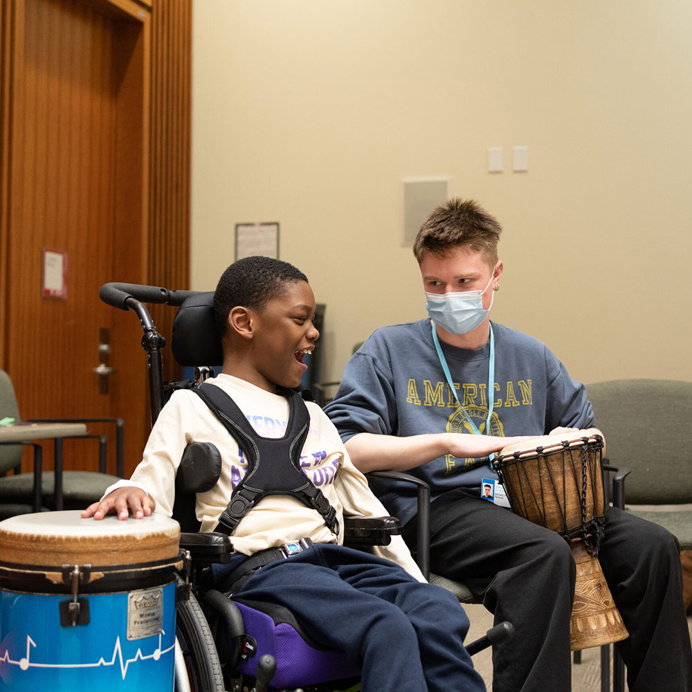 A young boy who uses a wheelchair smiling with an adult wearing a blue face mask. They're using drums.
