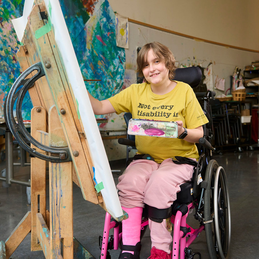 A teenage girl wearing a yellow t-shirt and using a pink wheelchair painting on a canvas in an art studio