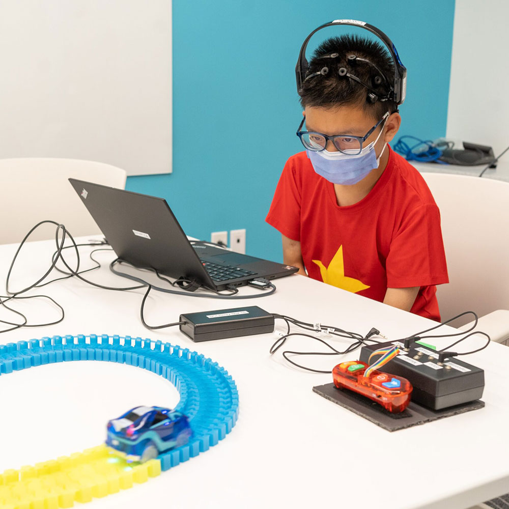 A young boy in a red t-shirt and wearing headphones sitting at a desk. There's a laptop in from of him as well as a toy car track.