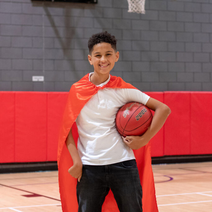 Zion on basketball court, holding basketball under one arm. Wearing his cape.