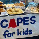 A bake sale table with a logo reading "Capes for Kids"