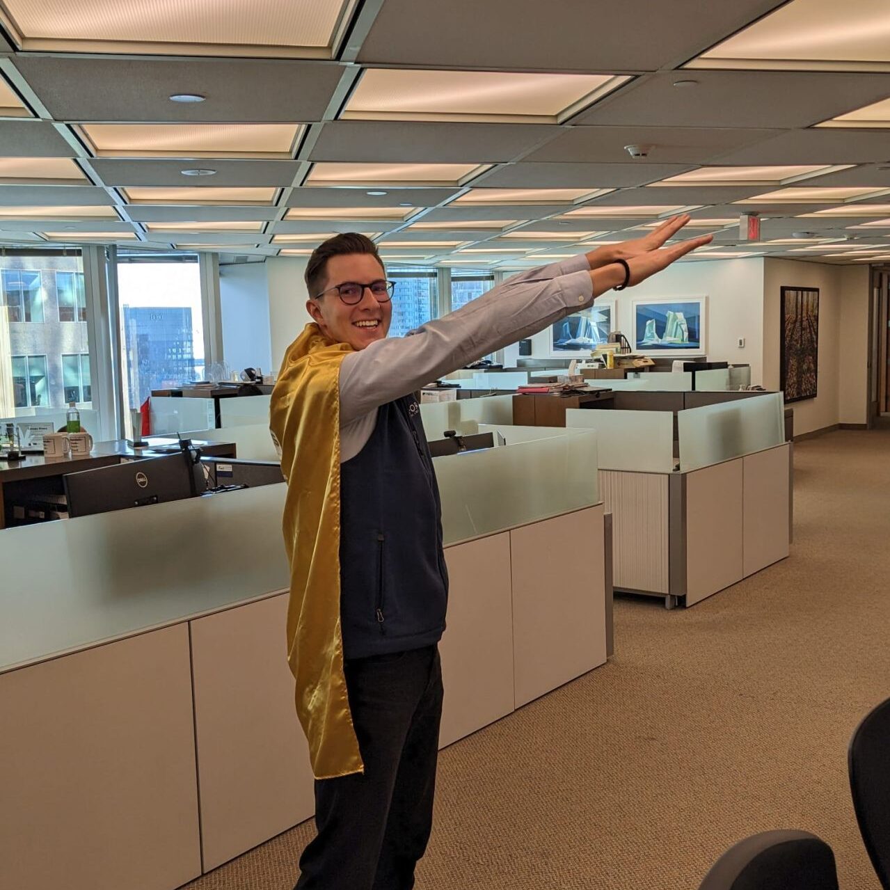 A smiling person wearing a gold cape and business wear standing in an office with their arms outstretched in a superhero pose