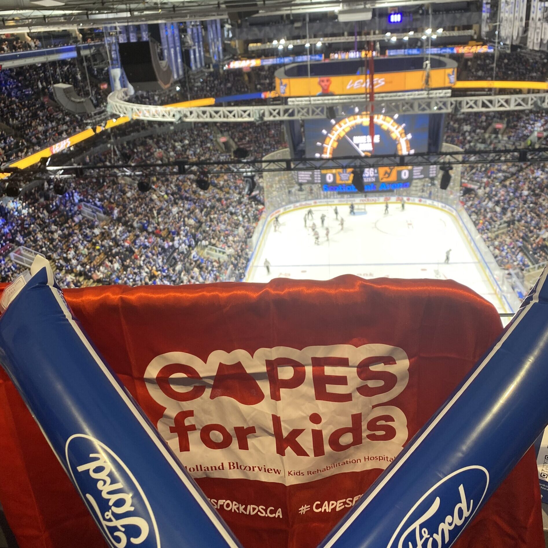 The view from a seat high up in a hockey area. A red cape with "Capes for Kids" logo and two blue noise maker sticks.