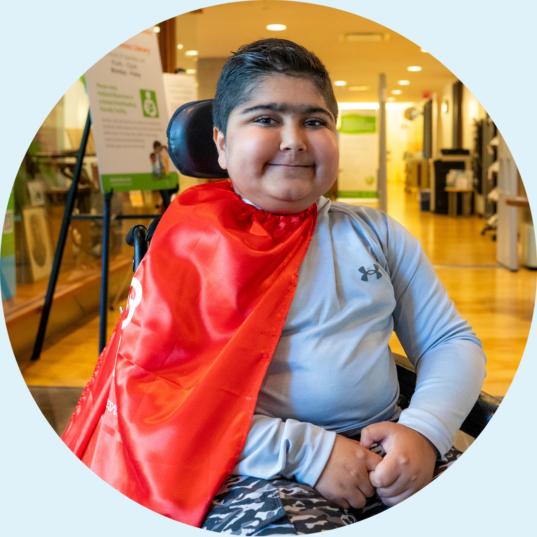 A young person who uses a wheelchair smiles and wears a red cape.