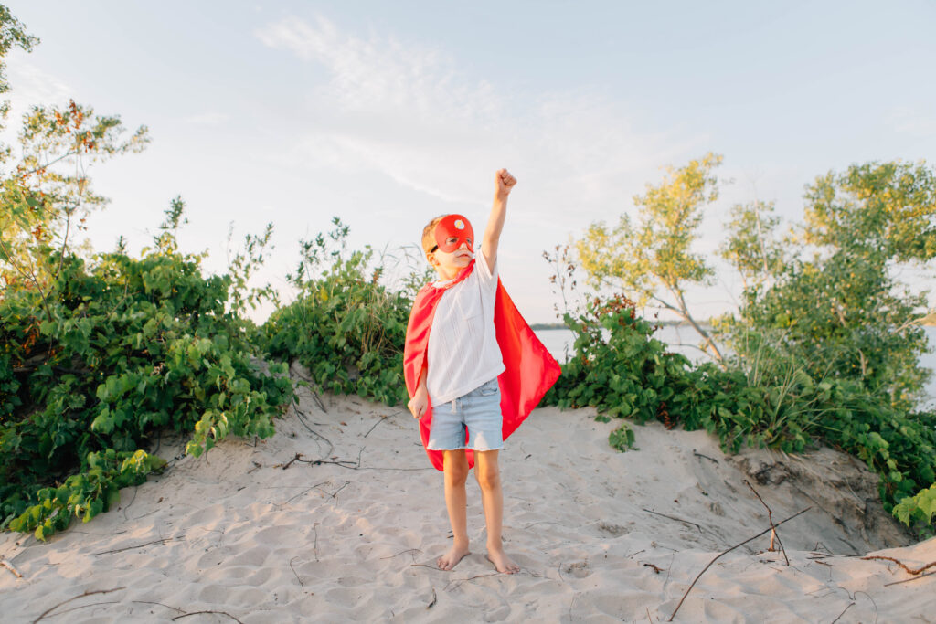 A young boy poses like a superhero on the beach wearing a cape and mask