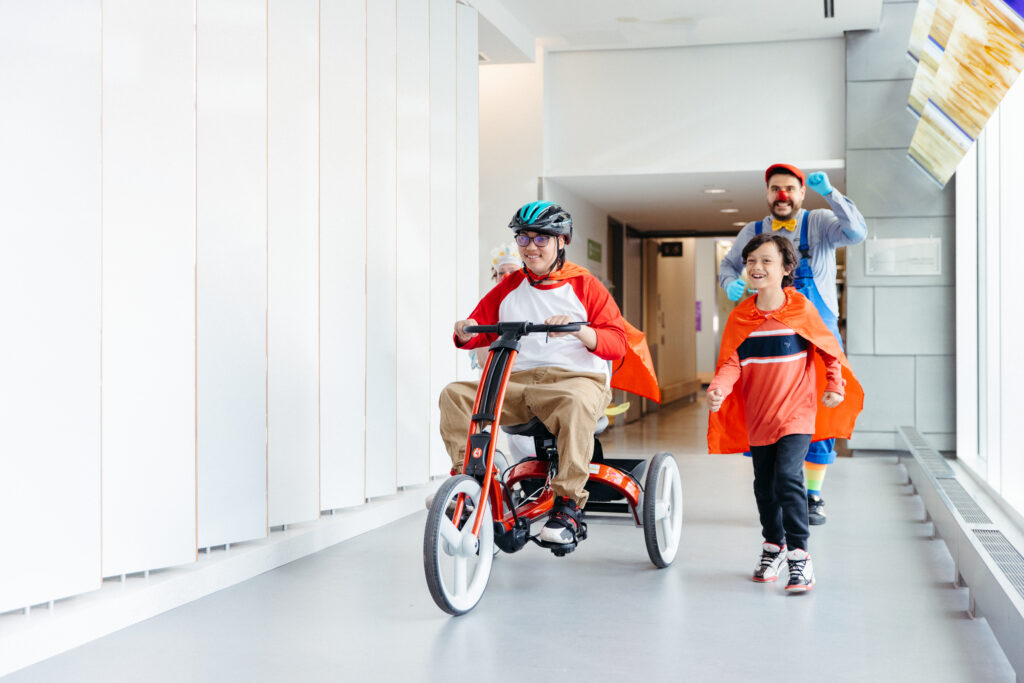 A therapeutic clown runs down the hospital hallways with two kids wearing red capes